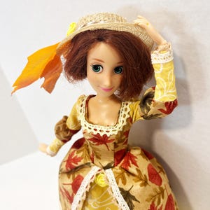 May include: A doll with auburn hair and green eyes wearing a tan dress featuring a red and brown autumn leaf pattern, a yellow skirt panel with white line drawings, and white lace trim. The doll wears a straw hat adorned with a yellow flower and leaf.