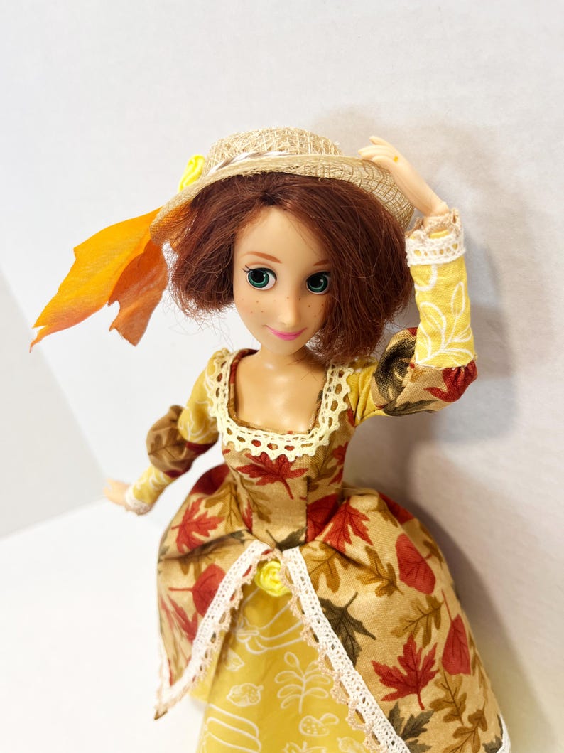 May include: A doll with auburn hair and green eyes wearing a tan dress featuring a red and brown autumn leaf pattern, a yellow skirt panel with white line drawings, and white lace trim. The doll wears a straw hat adorned with a yellow flower and leaf.