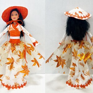 May include: A doll with long black hair wearing an orange wide-brimmed hat and a white dress featuring an orange bodice and a sheer skirt overlay decorated with metallic orange maple leaf patterns and a scalloped orange leaf-shaped hemline.