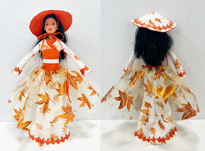 May include: A doll with long black hair wearing an orange wide-brimmed hat and a white dress featuring an orange bodice and a sheer skirt overlay decorated with metallic orange maple leaf patterns and a scalloped orange leaf-shaped hemline.