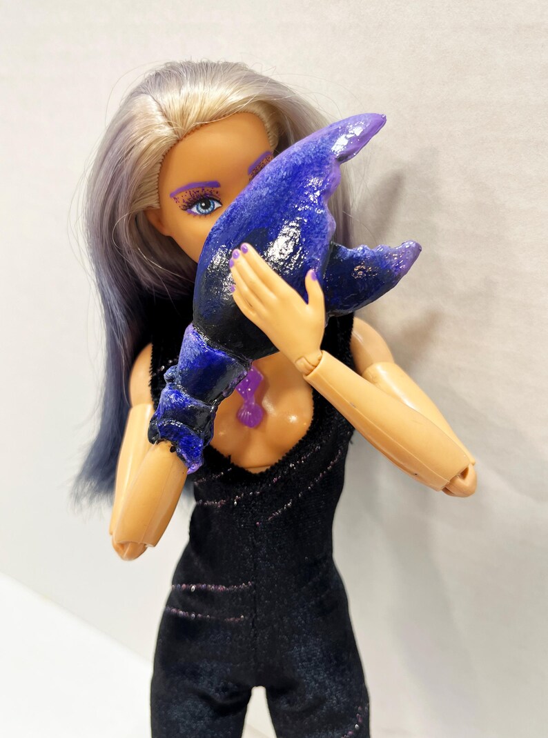 May include: A doll wearing a black jumpsuit with purple glitter accents, holding a large, blue, claw-shaped accessory. The doll has blue eye makeup and purple nail polish. The doll's hair is a mix of blonde and blue.