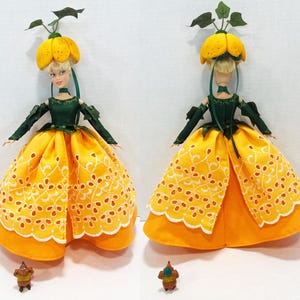 May include: A doll wearing a yellow pumpkin-shaped hat with green leaves, a dark green bodice with long sleeves, and a yellow skirt featuring white eyelet embroidery. A small brown figurine stands at the base of the doll in both views.
