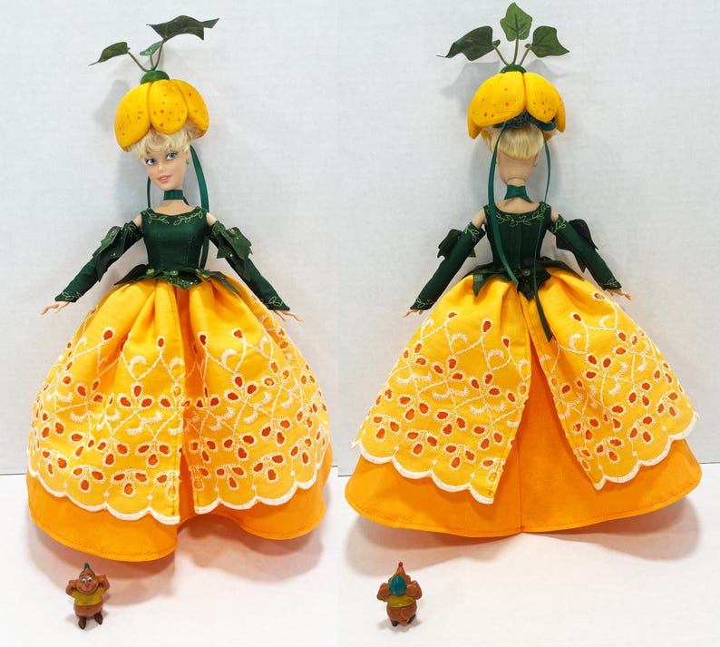 May include: A doll wearing a yellow pumpkin-shaped hat with green leaves, a dark green bodice with long sleeves, and a yellow skirt featuring white eyelet embroidery. A small brown figurine stands at the base of the doll in both views.