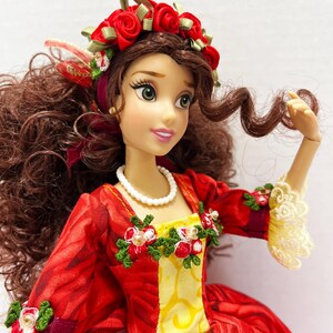 May include: A doll wearing a red and yellow dress with a floral design. The doll has brown hair and a red rose crown. The dress has white lace trim and a white beaded necklace. The doll's sleeves are adorned with floral accents.