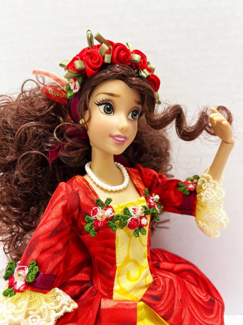 May include: A doll wearing a red and yellow dress with a floral design. The doll has brown hair and a red rose crown. The dress has white lace trim and a white beaded necklace. The doll's sleeves are adorned with floral accents.