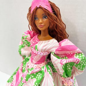 May include: A doll with brown hair and a pink crystal crown. The doll wears a dress with a pink, white, and green floral pattern. The dress has puffy sleeves and is adorned with pink sequins.