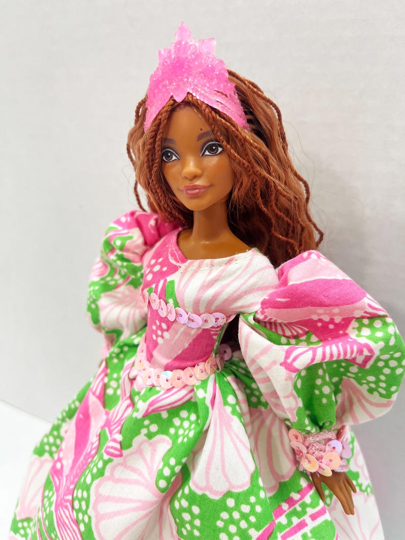 May include: A doll with brown hair and a pink crystal crown. The doll wears a dress with a pink, white, and green floral pattern. The dress has puffy sleeves and is adorned with pink sequins.