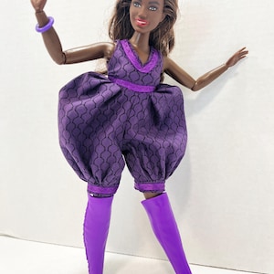 May include: A doll in a purple jumpsuit with a geometric pattern and matching knee-high boots. The jumpsuit features a V-neck and is trimmed with darker purple lace. A purple bracelet is on the doll's arm.