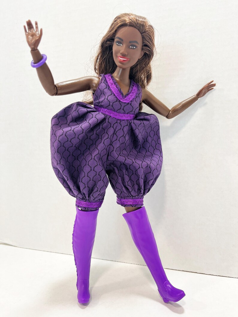 May include: A doll in a purple jumpsuit with a geometric pattern and matching knee-high boots. The jumpsuit features a V-neck and is trimmed with darker purple lace. A purple bracelet is on the doll's arm.
