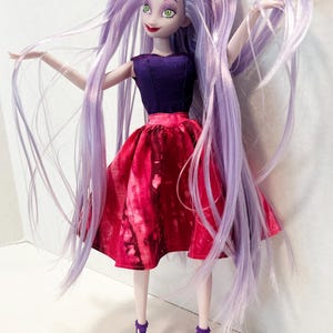 Pu&ograve; includere: Una bambola con lunghi capelli lisci color lavanda, occhi verdi e labbra rosse. La bambola indossa un corpetto senza maniche viola scuro abbinato a una gonna rosso vivace con un motivo maculato. Il completo &egrave; completato da sandali viola con tacco.
