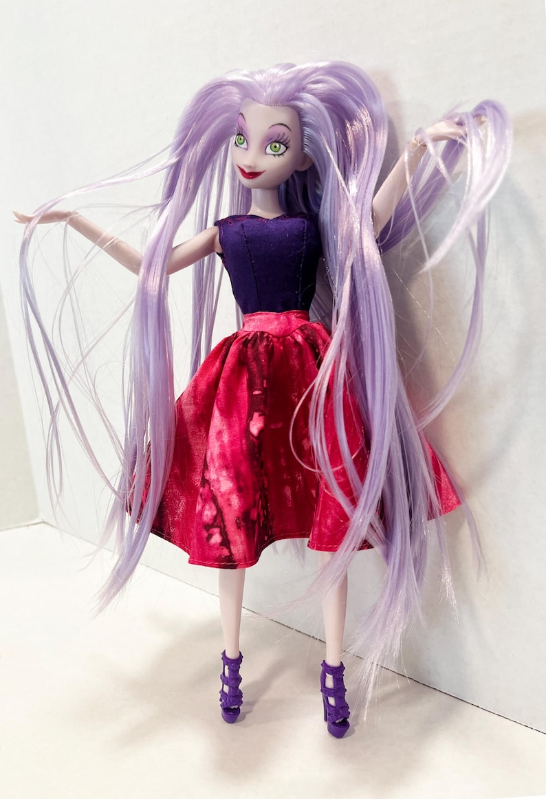 Pu&ograve; includere: Una bambola con lunghi capelli lisci color lavanda, occhi verdi e labbra rosse. La bambola indossa un corpetto senza maniche viola scuro abbinato a una gonna rosso vivace con un motivo maculato. Il completo &egrave; completato da sandali viola con tacco.