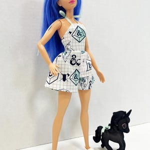 May include: A doll with long blue hair wearing a white halter-style romper featuring a grid pattern and Dungeons & Dragons themed icons including dice and weapons. The doll wears white shoes and stands next to a small black unicorn figurine.