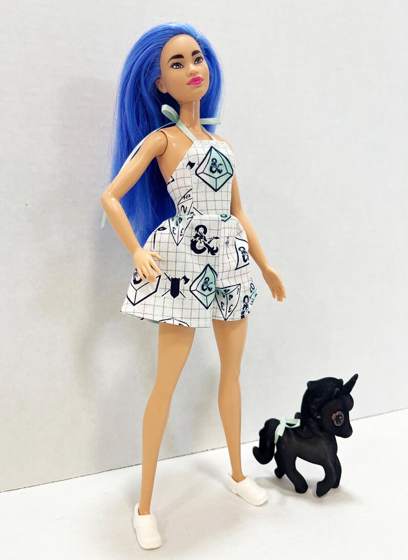 May include: A doll with long blue hair wearing a white halter-style romper featuring a grid pattern and Dungeons & Dragons themed icons including dice and weapons. The doll wears white shoes and stands next to a small black unicorn figurine.