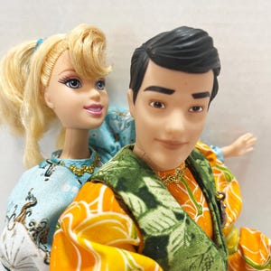 以下が含まれることがあります： 2体のファッションドール。金髪のドールは白と青の模様のライトブルーのドレスを着ています。男性のドールはオレンジ色のシャツと緑のベストを着ています。どちらのドールも金のネックレスをしています。