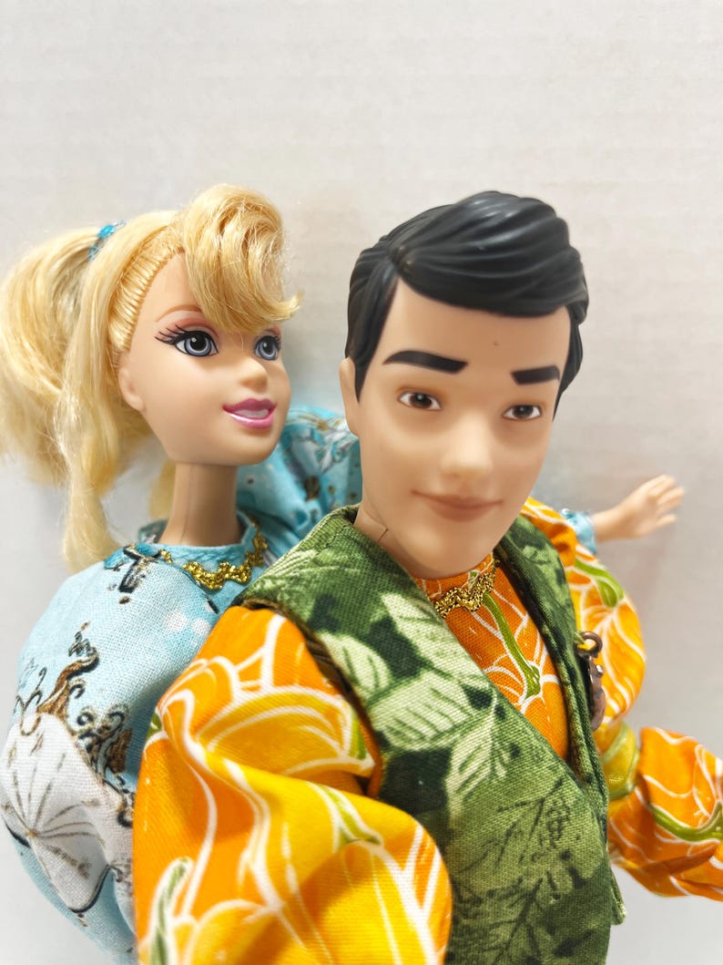 以下が含まれることがあります： 2体のファッションドール。金髪のドールは白と青の模様のライトブルーのドレスを着ています。男性のドールはオレンジ色のシャツと緑のベストを着ています。どちらのドールも金のネックレスをしています。
