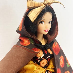 Op de afbeelding: Een pop met zwart haar draagt een bruine cape met capuchon met een rode appelprint en een beige geruite strik. De pop draagt een zwarte jurk met een patroon van appelschijfjes en een gele, gestructureerde rok, vastgemaakt met een zwarte sluiting.