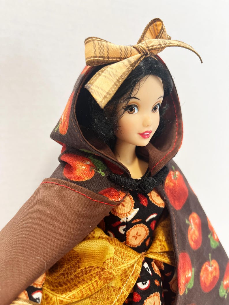 Op de afbeelding: Een pop met zwart haar draagt een bruine cape met capuchon met een rode appelprint en een beige geruite strik. De pop draagt een zwarte jurk met een patroon van appelschijfjes en een gele, gestructureerde rok, vastgemaakt met een zwarte sluiting.