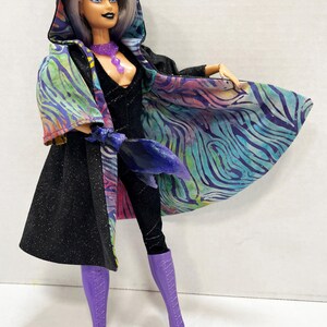 May include: A fashion doll in a black jumpsuit and purple boots, wearing a black hooded cape with a colorful, patterned lining. The doll has silver hair, purple eye makeup, and a purple necklace.