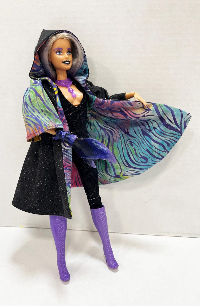May include: A fashion doll in a black jumpsuit and purple boots, wearing a black hooded cape with a colorful, patterned lining. The doll has silver hair, purple eye makeup, and a purple necklace.