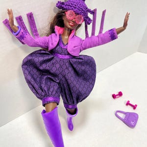May include: A doll dressed in a purple outfit, including a hat, glasses, jacket, and jumpsuit. The doll is wearing purple boots and has a matching purse and dumbbells. The hat has a pink and green pom-pom. The jumpsuit has a patterned design.