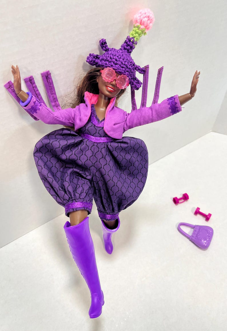May include: A doll dressed in a purple outfit, including a hat, glasses, jacket, and jumpsuit. The doll is wearing purple boots and has a matching purse and dumbbells. The hat has a pink and green pom-pom. The jumpsuit has a patterned design.