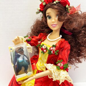 May include: A doll in a red rose-patterned dress with a yellow bodice, a red rose crown, and a pearl necklace. The doll is holding a small plastic box with a figure inside. The doll's dress has lace trim and floral accents.