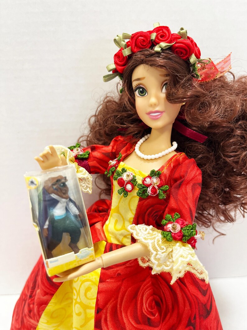 May include: A doll in a red rose-patterned dress with a yellow bodice, a red rose crown, and a pearl necklace. The doll is holding a small plastic box with a figure inside. The doll's dress has lace trim and floral accents.