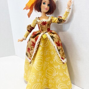 May include: A doll with auburn hair wearing a tan hat with an orange ribbon and a dress featuring a bodice with a red maple leaf print and a yellow skirt with a white line-art pattern of pumpkins, leaves, and mushrooms, accented with lace trim.