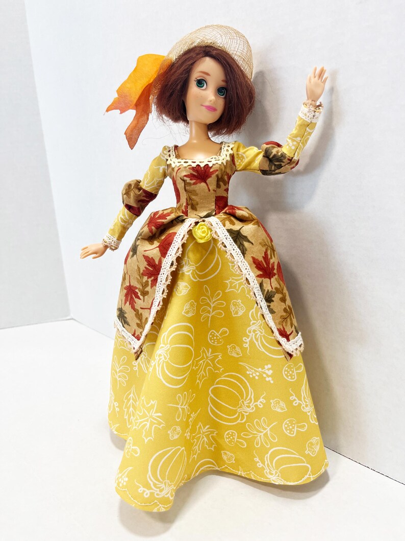 May include: A doll with auburn hair wearing a tan hat with an orange ribbon and a dress featuring a bodice with a red maple leaf print and a yellow skirt with a white line-art pattern of pumpkins, leaves, and mushrooms, accented with lace trim.