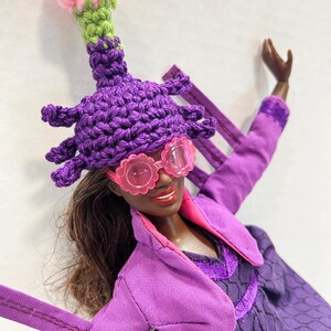 May include: A doll wearing a purple crochet hat with a pink and green top, pink flower-shaped sunglasses, and a purple outfit. The doll has brown hair and is smiling. The outfit includes a purple jacket and dress.