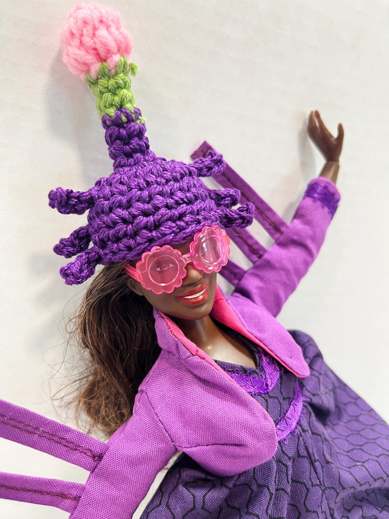 May include: A doll wearing a purple crochet hat with a pink and green top, pink flower-shaped sunglasses, and a purple outfit. The doll has brown hair and is smiling. The outfit includes a purple jacket and dress.