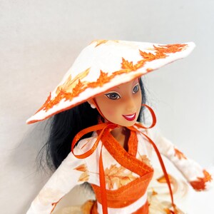 May include: A doll with long black hair wearing a white conical hat decorated with orange leaf patterns. The doll wears a matching white long-sleeved top with orange leaf prints, a wide orange waistband, and a full skirt with orange leaf accents.