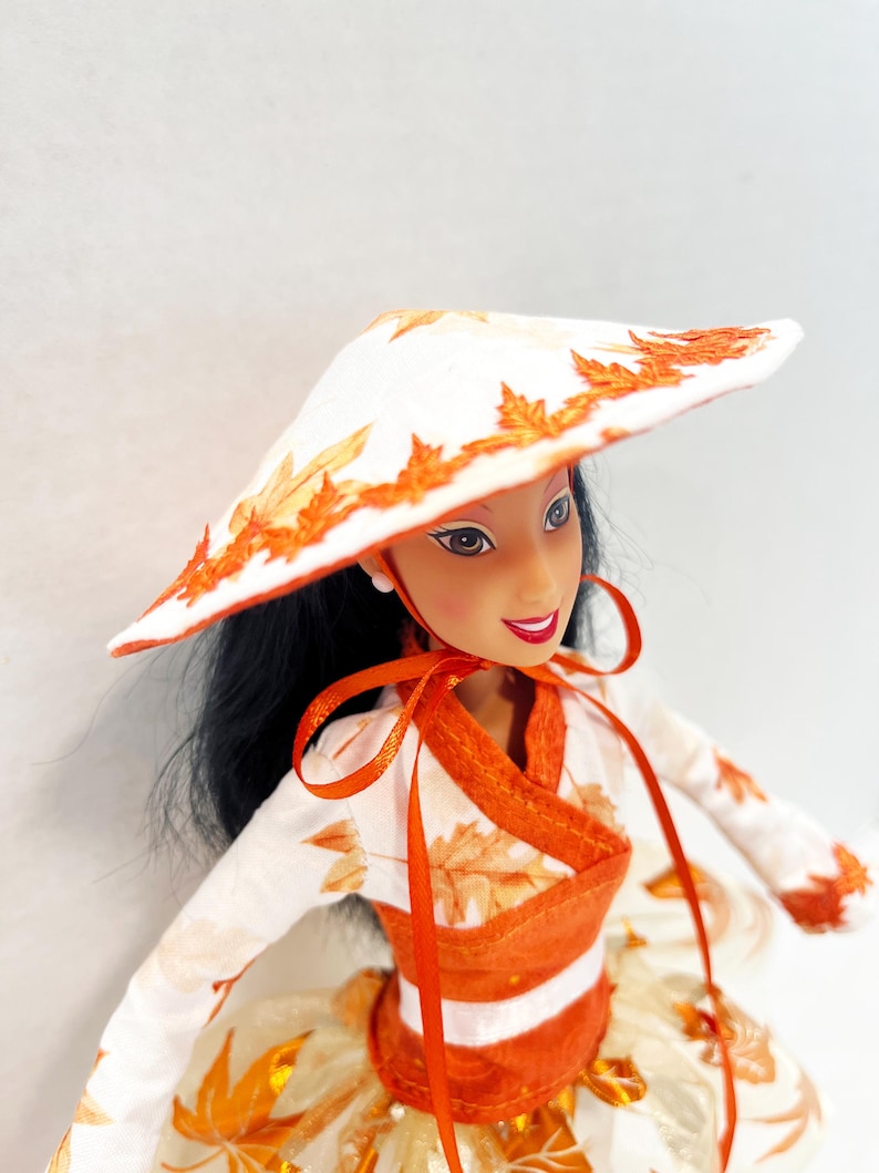 May include: A doll with long black hair wearing a white conical hat decorated with orange leaf patterns. The doll wears a matching white long-sleeved top with orange leaf prints, a wide orange waistband, and a full skirt with orange leaf accents.