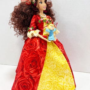 May include: A doll in a red rose-patterned gown with a yellow panel, holding a small blue angel figurine. The doll has brown curly hair, a floral crown, and a red and yellow bodice. The doll is wearing a pearl necklace.