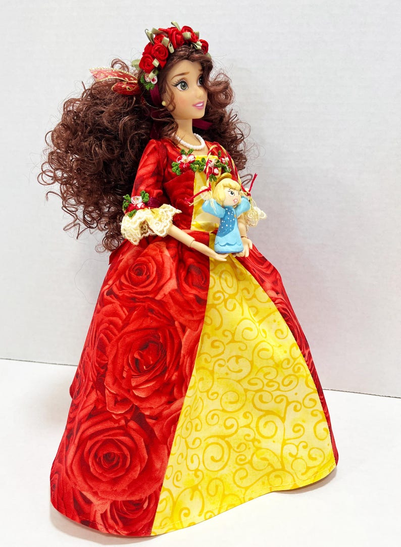 May include: A doll in a red rose-patterned gown with a yellow panel, holding a small blue angel figurine. The doll has brown curly hair, a floral crown, and a red and yellow bodice. The doll is wearing a pearl necklace.