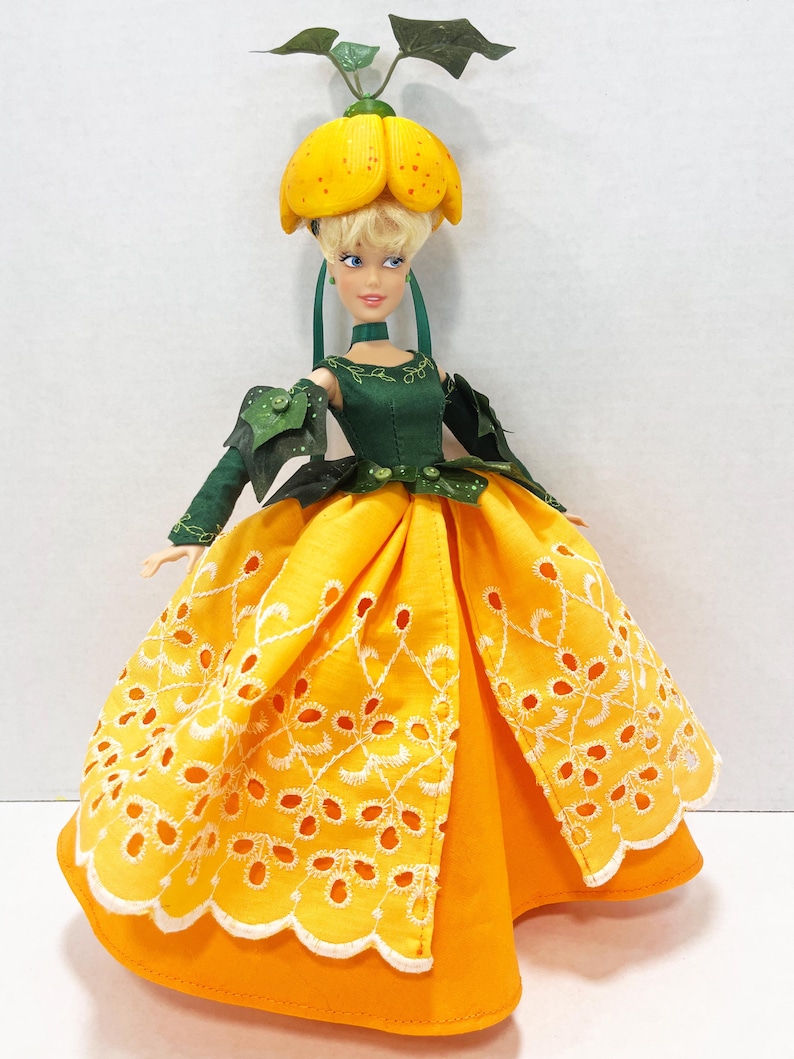 May include: A doll with blonde hair wearing a yellow flower-shaped headpiece with green leaves, a dark green bodice with long sleeves, and a bright yellow skirt featuring white eyelet embroidery patterns along the hem and front panels.