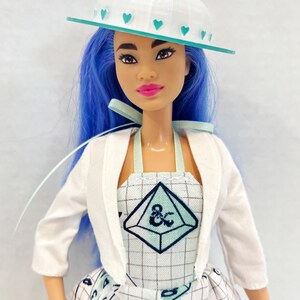 May include: A doll with bright blue hair wearing a white hat with teal heart accents, a white cropped jacket, and a white dress featuring a grid pattern, teal geometric shapes, and black ampersand symbols. The doll has dark eyes and pink lips.