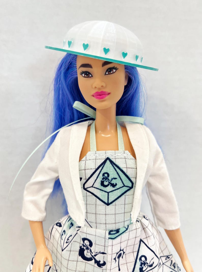 May include: A doll with bright blue hair wearing a white hat with teal heart accents, a white cropped jacket, and a white dress featuring a grid pattern, teal geometric shapes, and black ampersand symbols. The doll has dark eyes and pink lips.