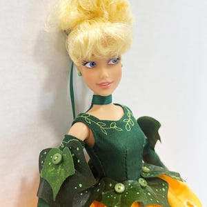 May include: A doll with blonde hair styled in an updo, blue eyes, and a green choker. The doll wears a dark green bodice with embroidered leaf details, green arm sleeves with leaf accents, and a bright yellow skirt with white eyelet trim.