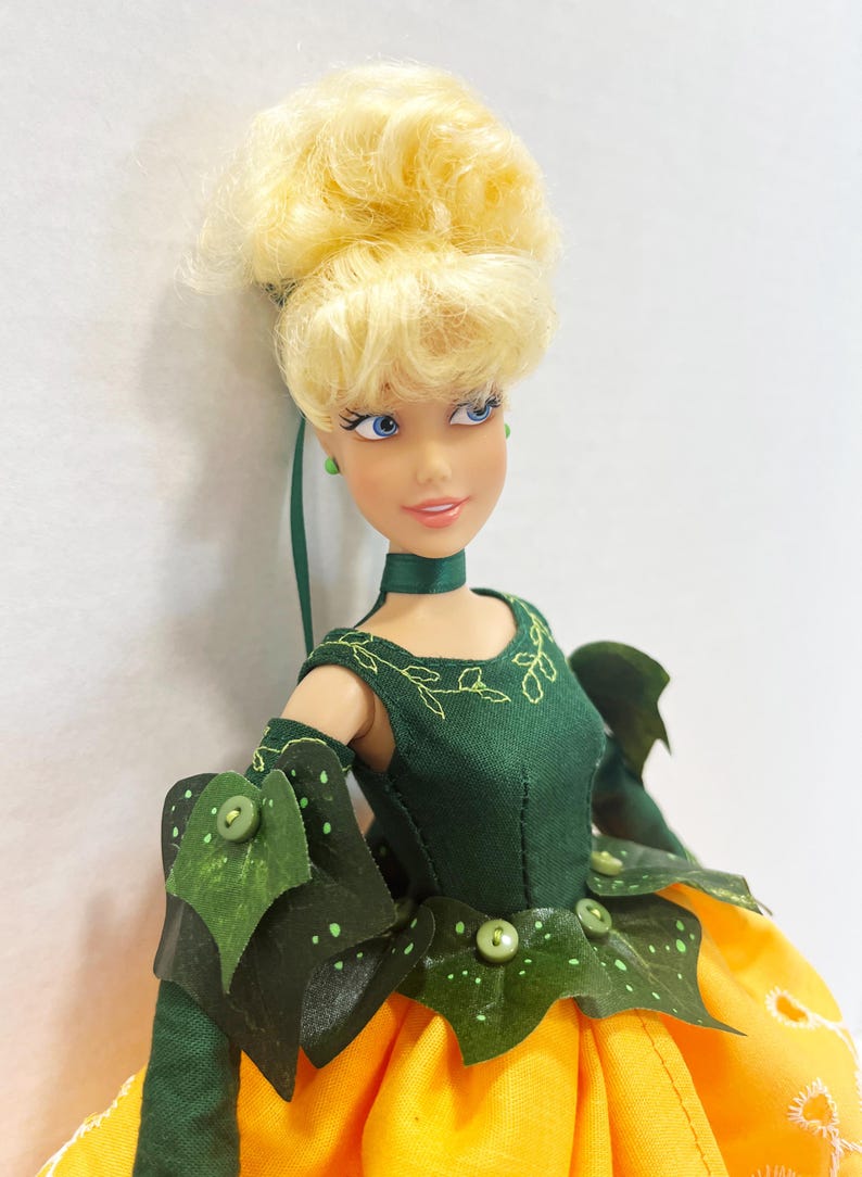May include: A doll with blonde hair styled in an updo, blue eyes, and a green choker. The doll wears a dark green bodice with embroidered leaf details, green arm sleeves with leaf accents, and a bright yellow skirt with white eyelet trim.