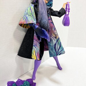 May include: A doll wearing a black hooded coat with a colorful zebra print design, purple boots, and sunglasses. The doll is holding a small purple purse and there is a purple flower accessory on the ground.
