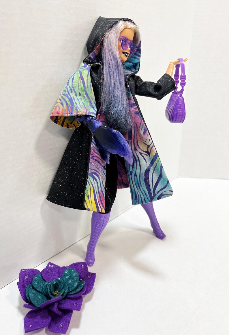 May include: A doll wearing a black hooded coat with a colorful zebra print design, purple boots, and sunglasses. The doll is holding a small purple purse and there is a purple flower accessory on the ground.
