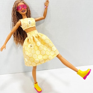 Puede incluir: Mu&ntilde;eca con un conjunto amarillo de dos piezas con estampado de panal y detalles de abeja. La mu&ntilde;eca tiene gafas de sol rosas, botas amarillas con suelas rosas y una pulsera rosa. Pelo largo casta&ntilde;o.