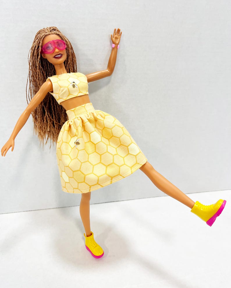 Puede incluir: Mu&ntilde;eca con un conjunto amarillo de dos piezas con estampado de panal y detalles de abeja. La mu&ntilde;eca tiene gafas de sol rosas, botas amarillas con suelas rosas y una pulsera rosa. Pelo largo casta&ntilde;o.