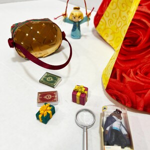 May include: A collection of miniature decorative items, including a woven basket with a burgundy handle, small gift boxes with yellow bows, and a magnifying glass. Also present are two miniature books, a figurine of a girl, and a figurine of a character in a clear plastic case.