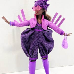 May include: A doll dressed in a purple outfit, featuring a jacket, jumpsuit, and boots. The doll wears a distinctive hat, pink sunglasses, and carries a matching handbag. The jumpsuit has a geometric pattern. The doll's ensemble is a vibrant display of purple.