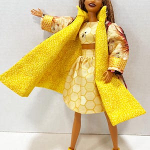 Puede incluir: Una mu&ntilde;eca vestida con un atuendo amarillo con un tema de abeja. El atuendo incluye un top corto, una falda y un abrigo largo. El abrigo tiene un estampado floral en las mangas y el cuello. La mu&ntilde;eca tambi&eacute;n lleva botas amarillas con suelas rosas.