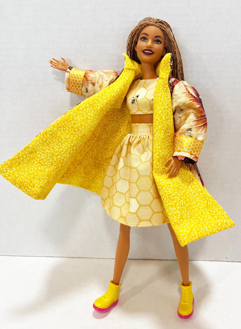 Puede incluir: Una mu&ntilde;eca vestida con un atuendo amarillo con un tema de abeja. El atuendo incluye un top corto, una falda y un abrigo largo. El abrigo tiene un estampado floral en las mangas y el cuello. La mu&ntilde;eca tambi&eacute;n lleva botas amarillas con suelas rosas.