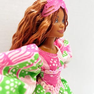 May include: A doll with brown skin and long, braided auburn hair, wearing a pink and green floral dress with puff sleeves. The dress is embellished with pink sequins. A pink, translucent headpiece adorns the doll's head.