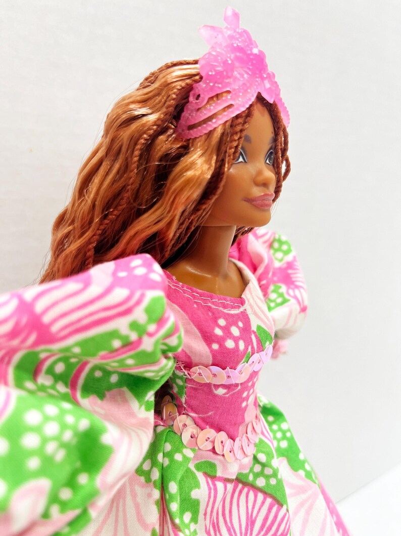 May include: A doll with brown skin and long, braided auburn hair, wearing a pink and green floral dress with puff sleeves. The dress is embellished with pink sequins. A pink, translucent headpiece adorns the doll's head.